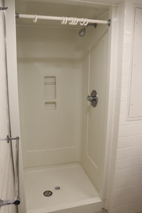 A clean, white shower stall with a single showerhead, a soap shelf, and a towel bar on the outside wall. The shower curtain rod is empty, except for several white curtain rings. The walls are tiled, and the stall features a simple, functional design.
